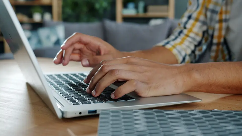 Foto ilustrativa: mãos digitando num notebook numa mesa de casa